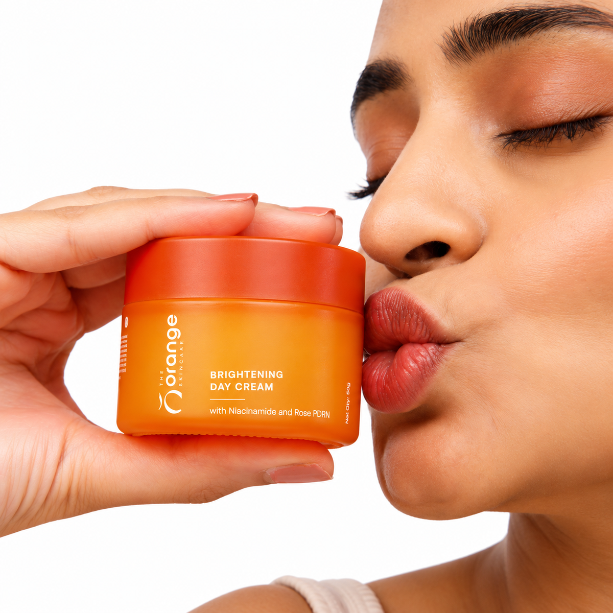 A close-up lifestyle shot of a woman holding an orange cosmetic jar next to her lips, with the brand name 'The Orange' and product name 'Brightening Day Cream' visible on the jar.