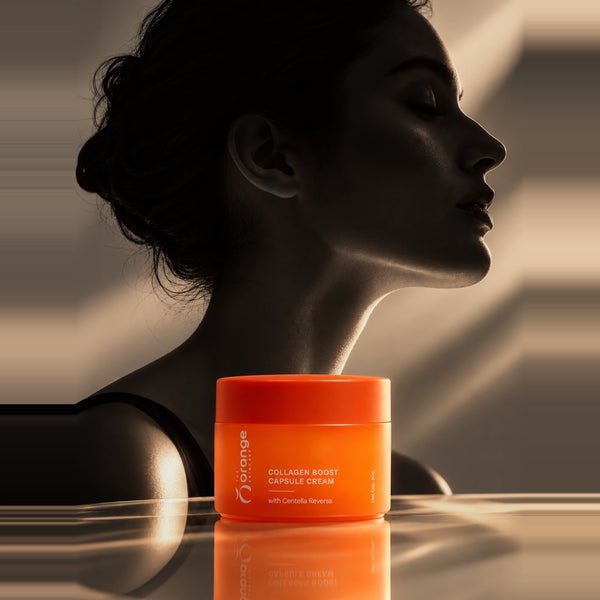 An orange cosmetic jar labeled 'orange' and 'Collagen Boost Capsule Cream' placed on a reflective surface in front of a woman's face in warm, dramatic lighting.