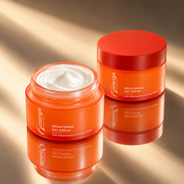 Two orange cosmetic jars of Orange Beauty Brightening Day Cream, one open showing white cream and one closed, on a warm beige reflective surface with soft shadows.